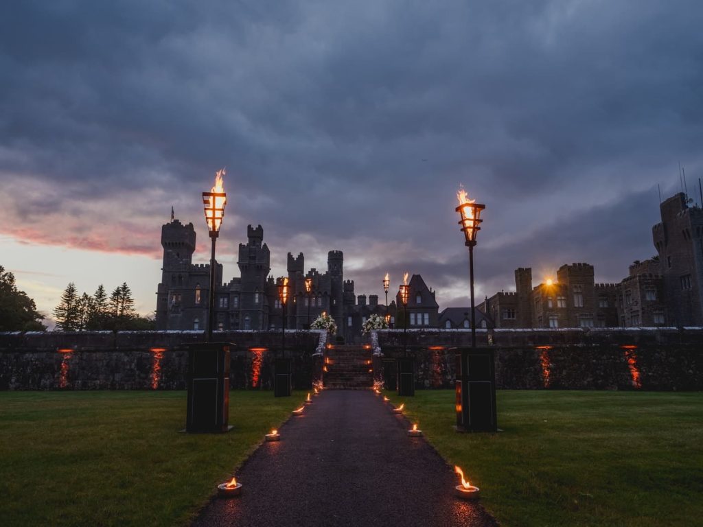 Castle wedding in Ireland at Ashford Castle, County Mayo — illuminated pathway leading to the castle at nightfall, with fire lamps lining the entrance and the estate visible in the background.