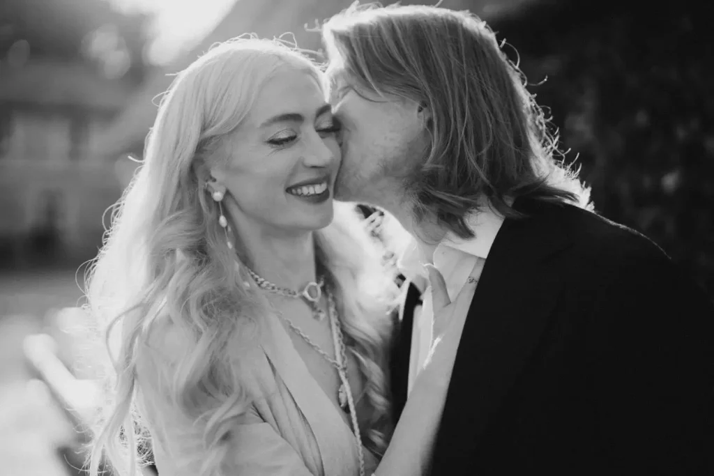 Black-and-white photo of a smiling woman in a wedding dress being kissed on the cheek by a man in a dark suit outdoors.