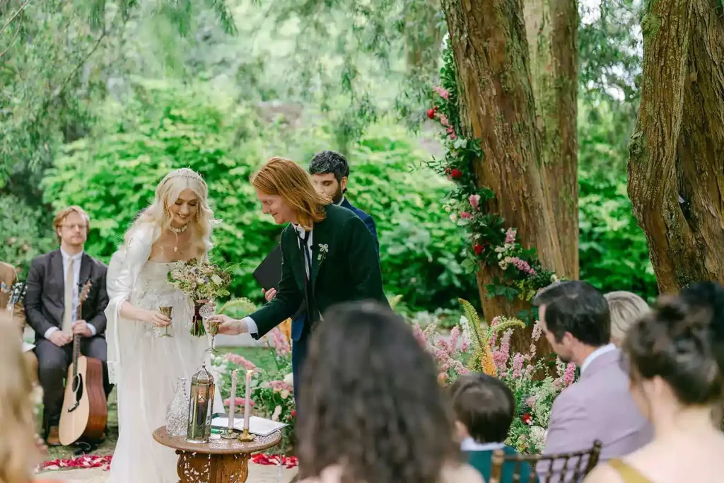 Bride in a white gown and groom in a dark suit raise glasses during a garden wedding under a floral arch, with guests seated nearby.