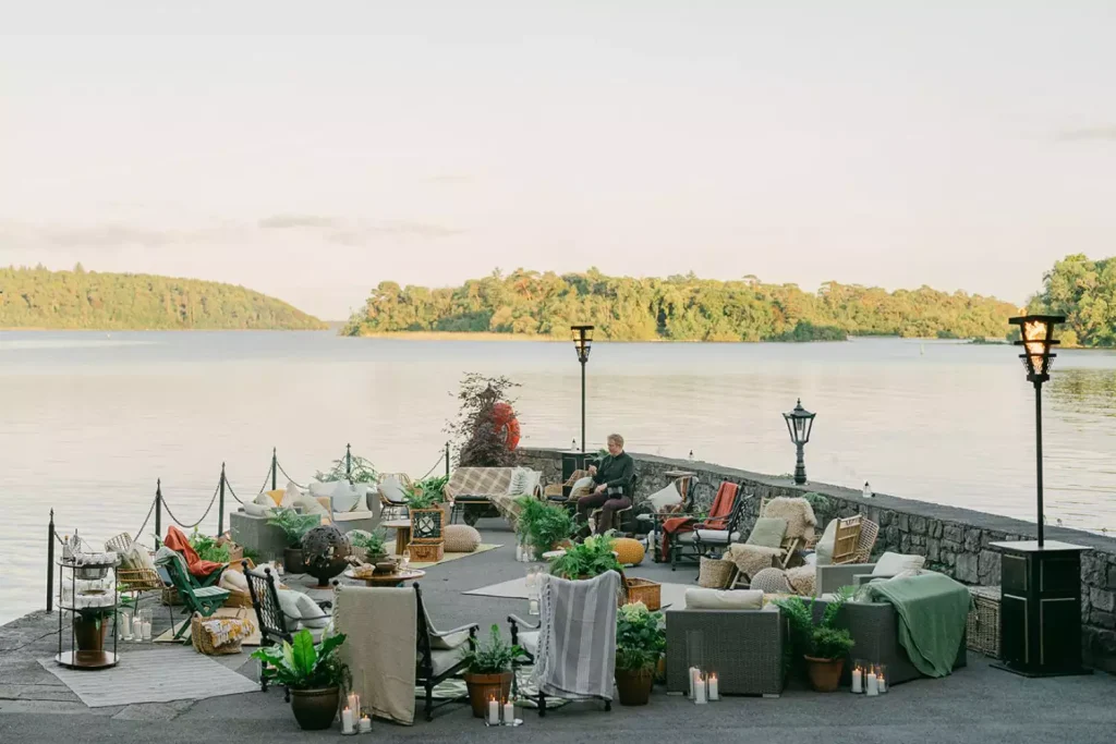 Destination wedding in Ireland at Ashford Castle, County Mayo — outdoor dock setup overlooking the lake with lounge furniture, cushions, candles, greenery, and a musician seated ready to perform.