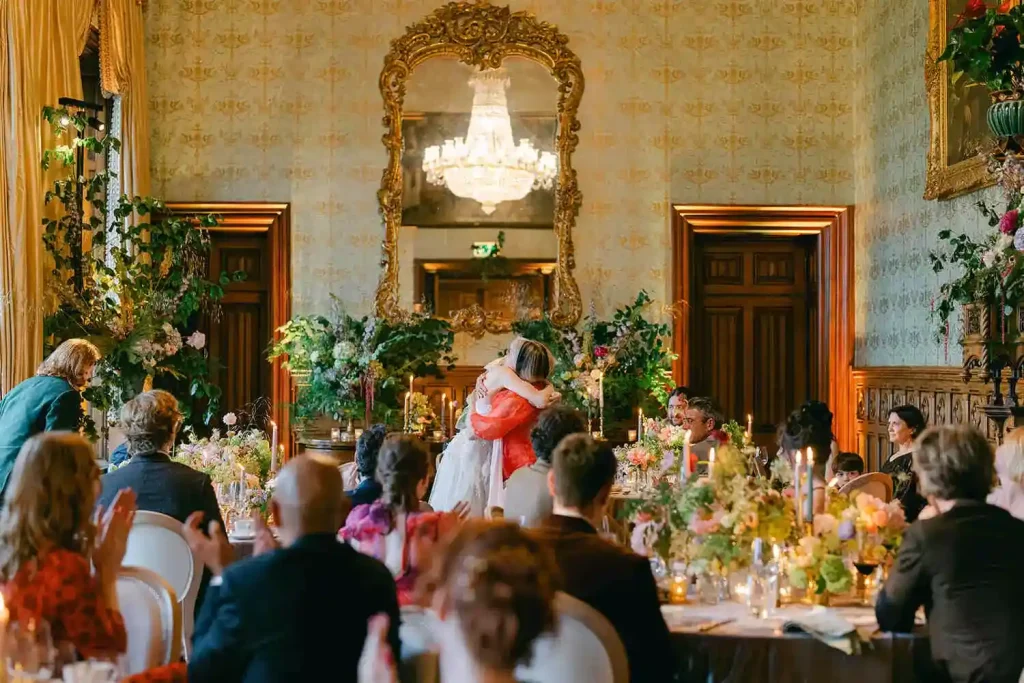 Bride and groom hug at a lavish wedding reception in an ornate gold-framed room with floral centerpieces