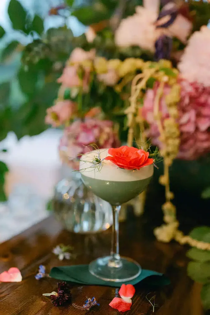 Cocktail glass with a red flower garnish on a wooden table, blurred colorful floral backdrop behind it.