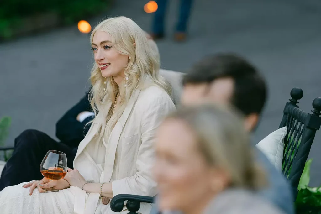 Blonde woman in a white outfit sits outdoors, smiling while holding a wine glass.