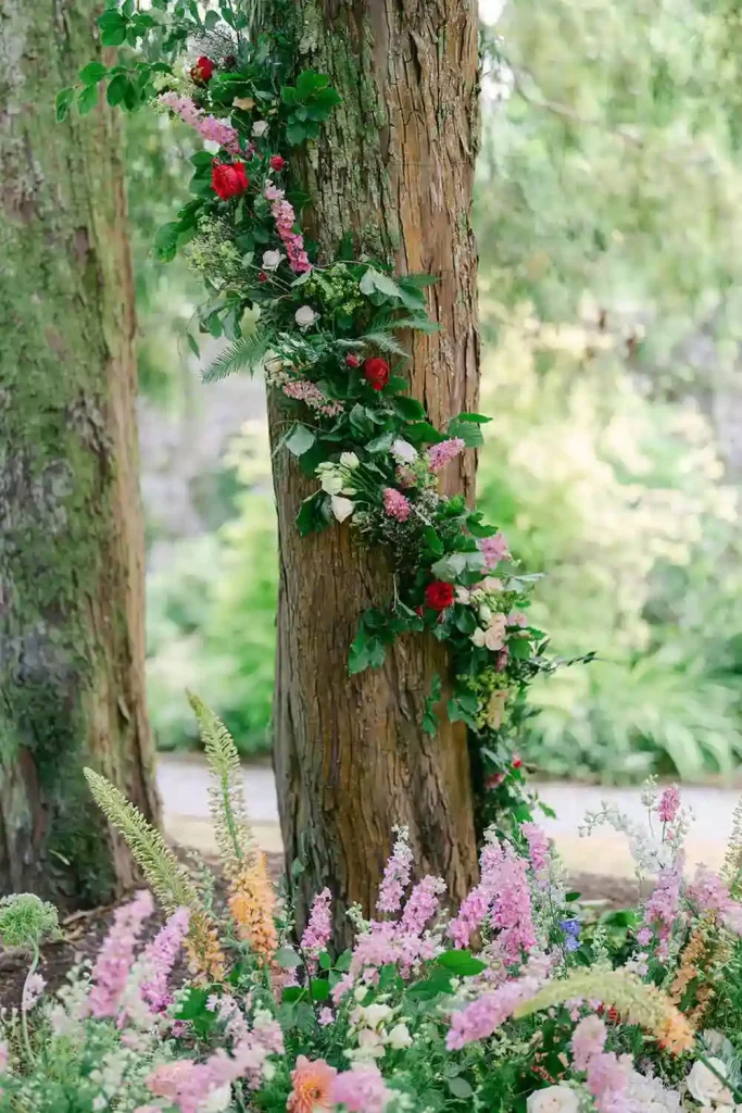 Destination wedding ceremony at Ashford Castle, County Mayo — floral installation at the base of a tree with flowers and greenery wrapped around the trunk for an outdoor ceremony setting.