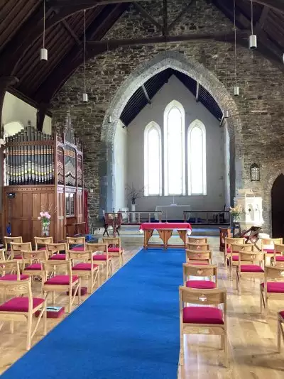 Stone church interior with a blue aisle runner leading to the altar; wooden chairs with red cushions, organ on the left, and tall arched windows behind the altar.