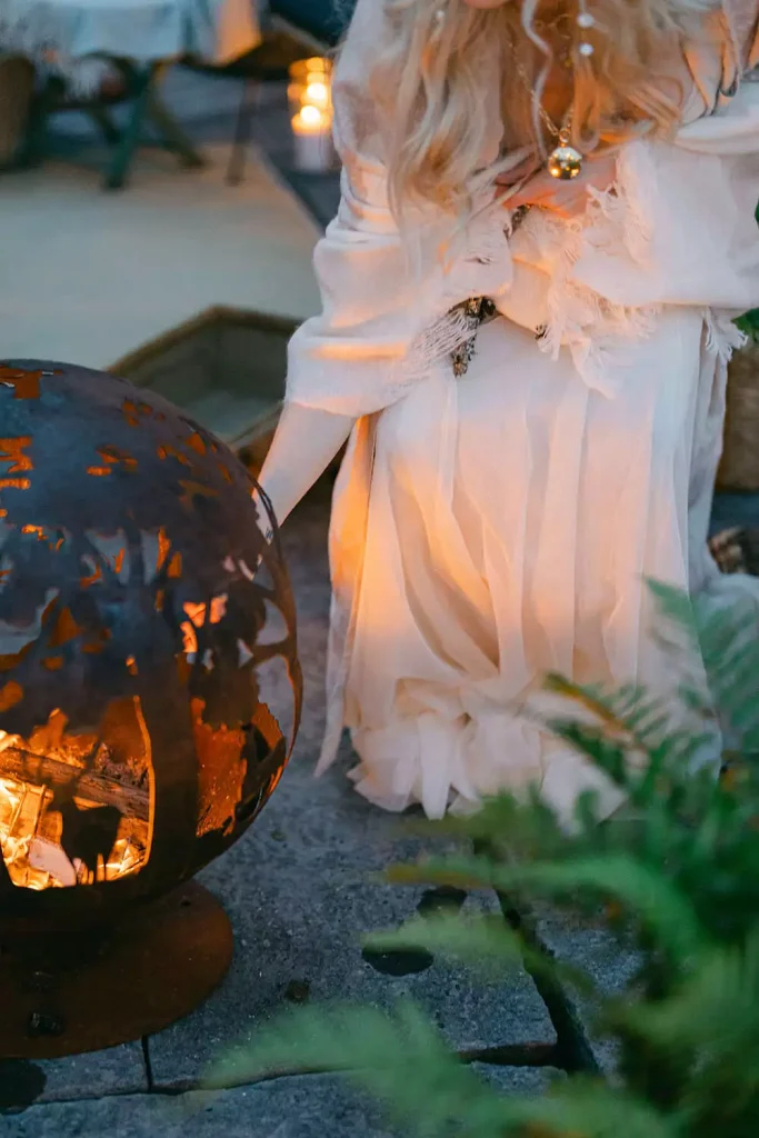 Person in a pale dress kneels beside a glowing carved pumpkin lantern outdoors at dusk.