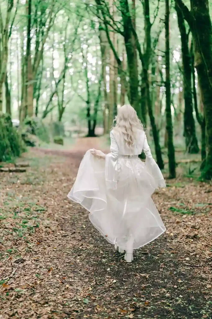 Woman in a white wedding dress walking away along a leaf-covered forest path.