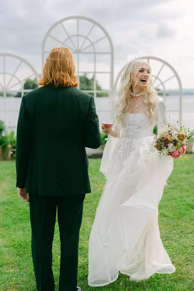 Bride in a white wedding gown with bouquet joyfully facing a man in a black suit outdoors, with arched white structures by a lake in the background.