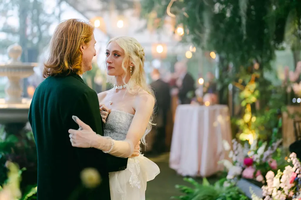 Bride in a lace strapless gown and pearl choker embracing her partner as they dance at a garden wedding, warm bokeh lights in the background.