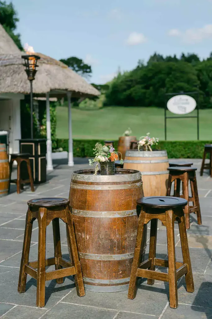 Outdoor patio with wooden wine-barrel tables and stools, floral centerpieces, and a thatched-roof shelter on a sunny day