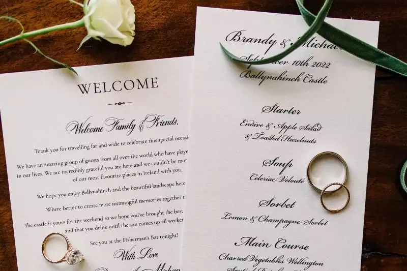 Wedding invitation cards on a wooden table with rings; left card begins with 'WELCOME' and a message, right card lists the menu and couple's names.