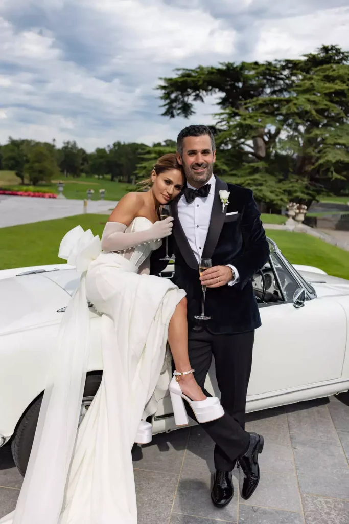 Irish destination wedding at Adare Manor, County Limerick — bride seated on a vintage white car with the groom standing beside her, both holding champagne glasses outdoors on the estate.