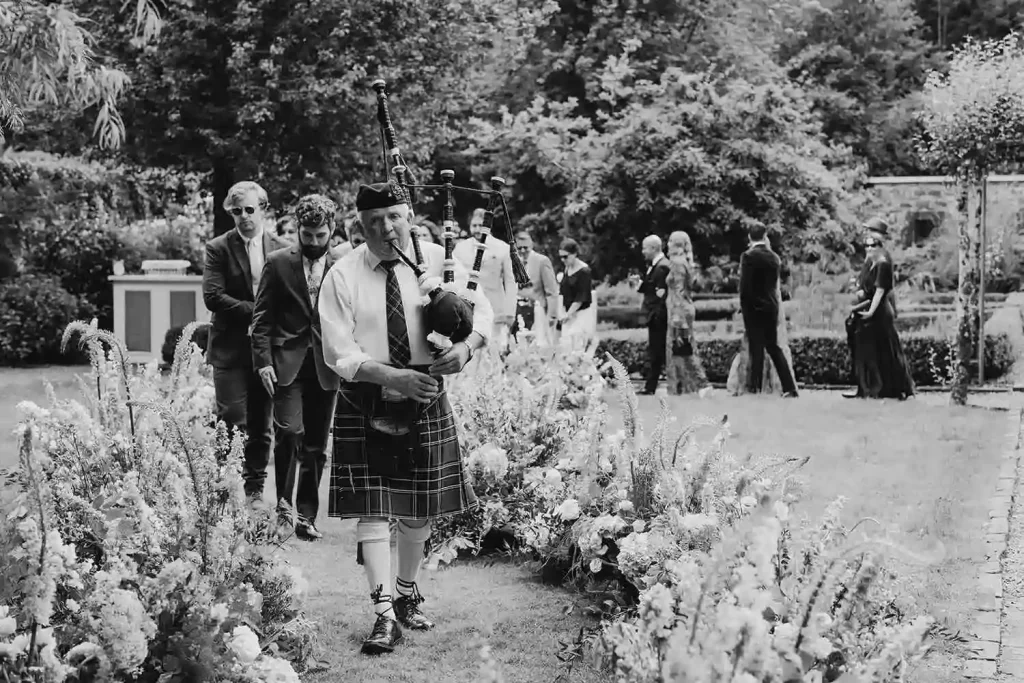 Irish destination wedding at Ashford Castle, County Mayo — bagpiper leading guests along a curved floral aisle at the conclusion of an outdoor ceremony beneath the estate trees.