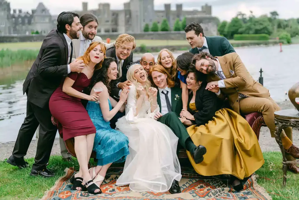 Group of friends and the wedding couple posing closely on a blanket by a lake during a celebration by the water.