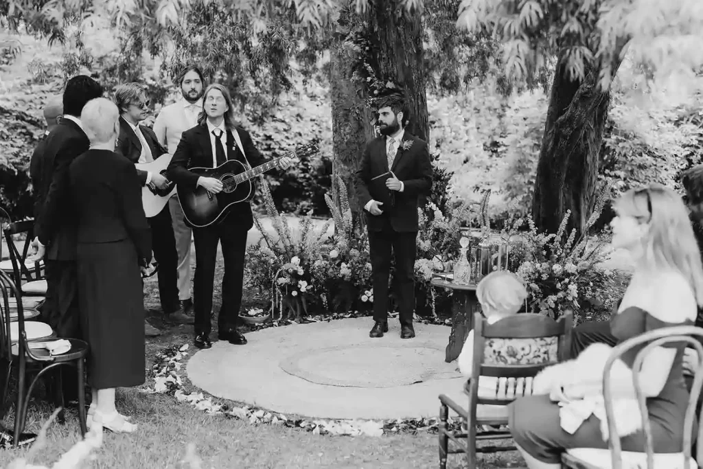 Irish destination wedding ceremony at Ashford Castle, County Mayo — groom standing at the altar performing with a guitar as guests listen during an outdoor ceremony beneath the estate trees.