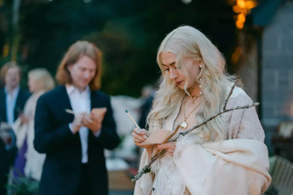 Blonde woman in a light shawl writing on a small card at an outdoor event, with people in formal attire blurred in the background.