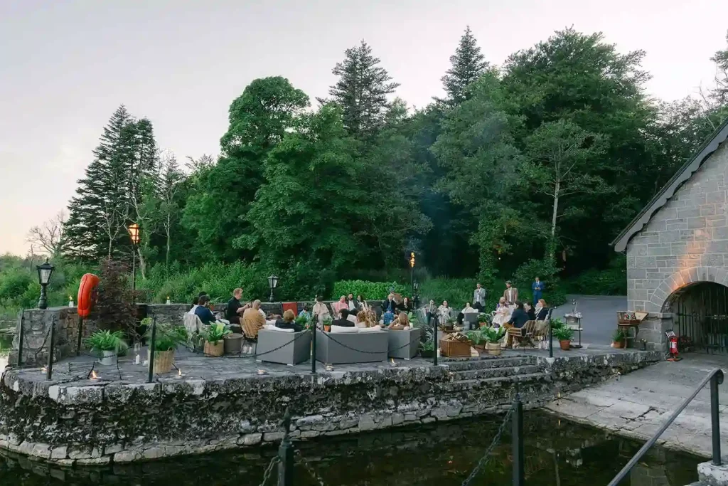 Destination wedding in Ireland at Ashford Castle, County Mayo — lakeside summer solstice gathering with guests relaxing on lounge seating during a live music performance.